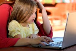 A little girl is sitting on her mother's lap while she is working on her laptop