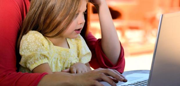 A little girl is sitting on her mother's lap while she is working on her laptop